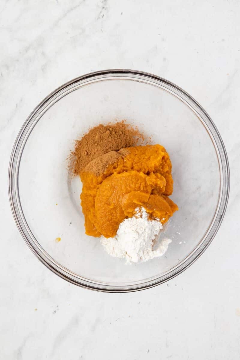 A glass bowl contains pumpkin puree, ground cinnamon, and flour on a white marble surface.