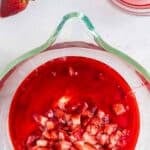 A glass bowl filled with chopped strawberries in red gelatin mixture sits on a white surface, with whole strawberries and a small dish of red liquid nearby.