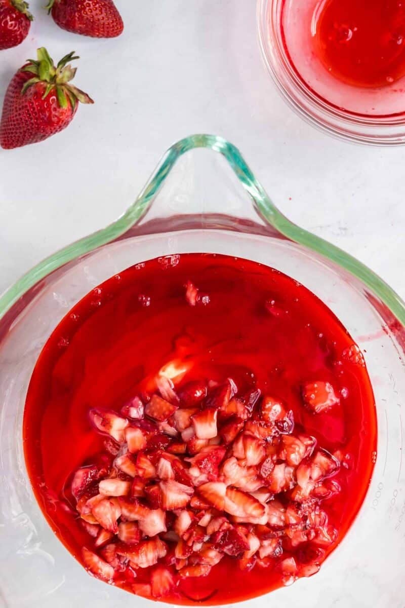 A glass bowl filled with chopped strawberries in red gelatin mixture sits on a white surface, with whole strawberries and a small dish of red liquid nearby.