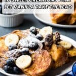 A plate of French toast topped with sliced bananas, blueberries, and powdered sugar sits on a table with another plate and a ramekin in the background.