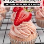A strawberry cupcake with pink frosting and half a strawberry on top sits on a cooling rack with other similar cupcakes in the background.