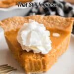 A slice of pumpkin pie with whipped cream on top sits on a white plate next to a fork, with another pie slice in the background.