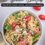 A bowl of pasta salad with bacon, cherry tomatoes, spinach, and shredded cheese, placed on a table with a gold fork and scattered tomatoes nearby.
