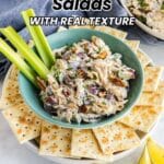 A bowl of chicken salad with celery sticks and crackers, garnished with lemon wedges, with text reading "16 Bold Chicken Salads With Real Texture.