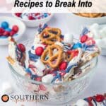 A glass bowl filled with white chocolate bark topped with pretzels, red and blue candies, and mini marshmallows, with more treats and ingredients in the background.
