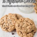 Two large chocolate chip oatmeal cookies on parchment paper with a glass of milk and a green striped straw, chocolate chips, and oats scattered nearby.