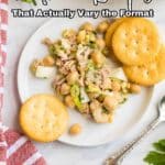 A plate with tuna salad, chickpeas, chopped celery, and three round crackers next to a fork, with a striped napkin and a bowl of salad in the background.