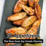 A slate serving board with seasoned chicken wings and a pickle spear, overlaid with the text "18 Appetizers That Keep Game Day Crowds Cheering.