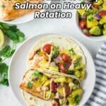 Two salmon tacos topped with avocado salsa and sauce on a plate, with a side of salsa and a partial plate of salmon in the background.