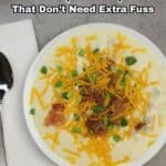 A bowl of creamy soup topped with shredded cheddar cheese, bacon, and green onions, placed on a white plate next to a spoon and napkin.