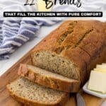 A loaf of sliced banana bread on a wooden cutting board with butter, a knife, and a striped kitchen towel nearby.