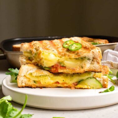 Two halves of a grilled cheese sandwich with melted cheese, sliced jalapeños, and cucumbers are stacked on a white plate—a delicious addition to flavorful vegetarian meals—set against a backdrop of a pan and kitchen towel.