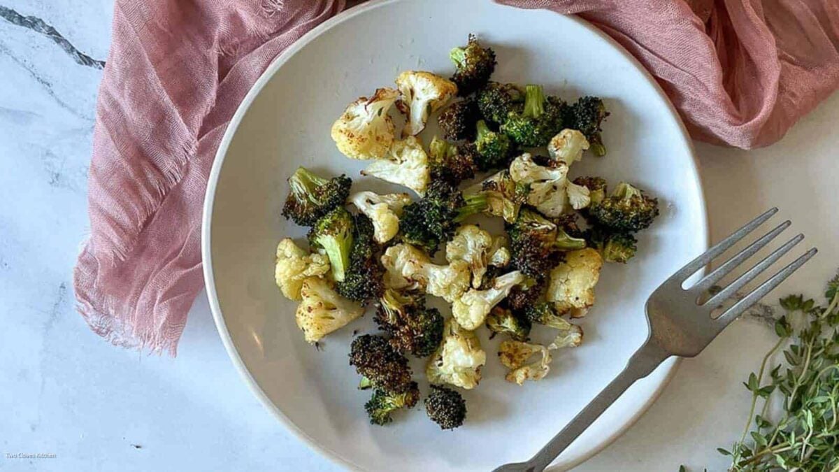 A white plate with roasted broccoli and cauliflower florets, a tasty addition to air fryer side dishes, is placed next to a metal fork and a pink cloth on a white surface.
