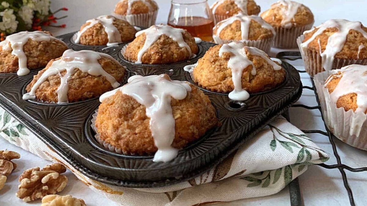 A tray of freshly baked muffins with white icing drizzled on top, surrounded by more muffins and a small glass jar on a patterned cloth, offers wholesome flavor with the delightful addition of crunchy walnuts.