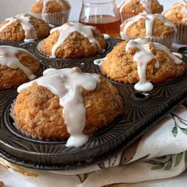A tray of freshly baked muffins with white icing drizzled on top, surrounded by more muffins and a small glass jar on a patterned cloth, offers wholesome flavor with the delightful addition of crunchy walnuts.