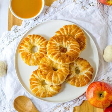 A plate of six round, golden-brown pastries with a soft donut texture is drizzled with icing and placed on a white cloth, surrounded by apples and a bowl of sauce.