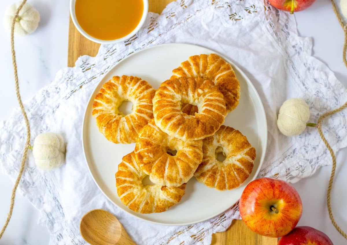 A plate of six round, golden-brown pastries with a soft donut texture is drizzled with icing and placed on a white cloth, surrounded by apples and a bowl of sauce.