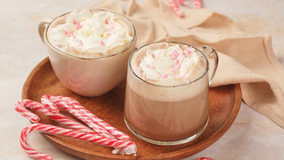 Two mugs of hot chocolate topped with whipped cream and crushed peppermint sit on a wooden plate with several candy canes, perfect for savoring during holiday cooking or adding to your favorite Christmas recipes.
