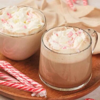 Two mugs of hot chocolate topped with whipped cream and crushed peppermint sit on a wooden plate with several candy canes, perfect for savoring during holiday cooking or adding to your favorite Christmas recipes.