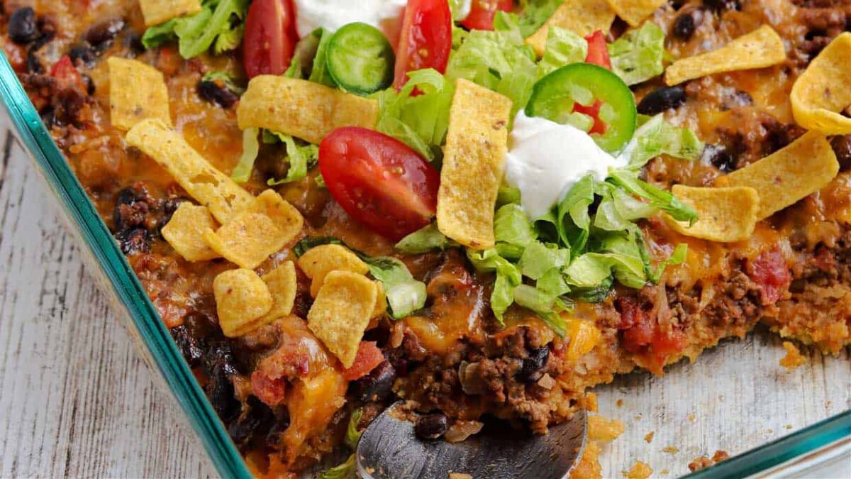 A glass dish of taco casserole topped with lettuce, tomato, jalapeño slices, sour cream, and corn chips—one spoonful missing—perfect for those seeking flavorful casseroles and new casserole ideas.