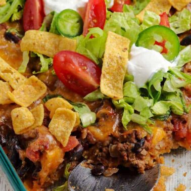 A glass dish of taco casserole topped with lettuce, tomato, jalapeño slices, sour cream, and corn chips—one spoonful missing—perfect for those seeking flavorful casseroles and new casserole ideas.