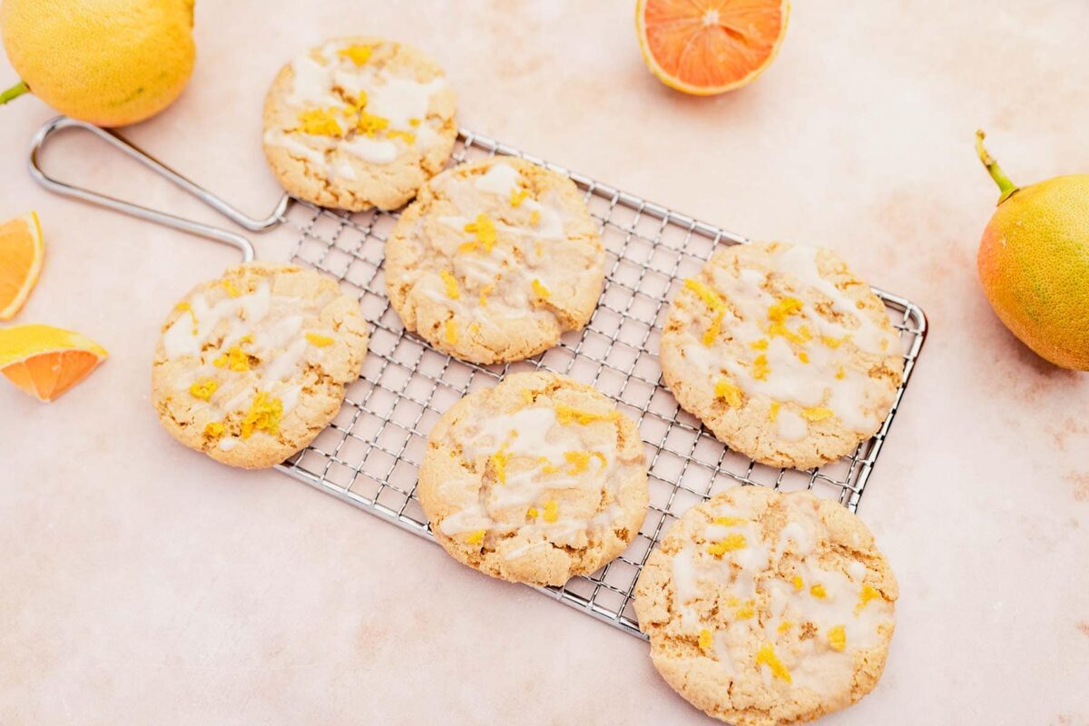 Six glazed citrus cookies on a cooling rack, surrounded by whole and sliced oranges and grapefruits&mdash;perfect gluten-free treats for anyone who loves zesty desserts.