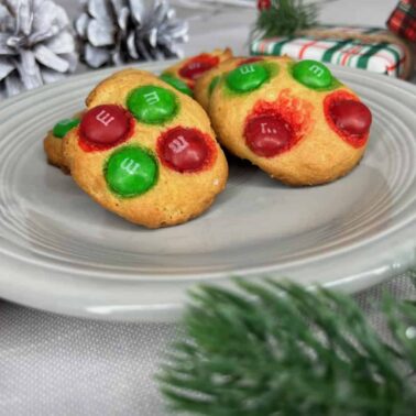 Two cookies topped with red and green M&Ms sit on a gray plate, surrounded by holiday table decorations like pine cones, greenery, and a wrapped giftโperfect for festive Christmas recipes.