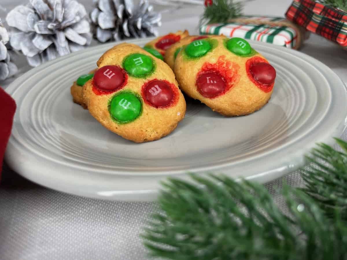 Two cookies topped with red and green M&Ms sit on a gray plate, surrounded by holiday table decorations like pine cones, greenery, and a wrapped gift—perfect for festive Christmas recipes.
