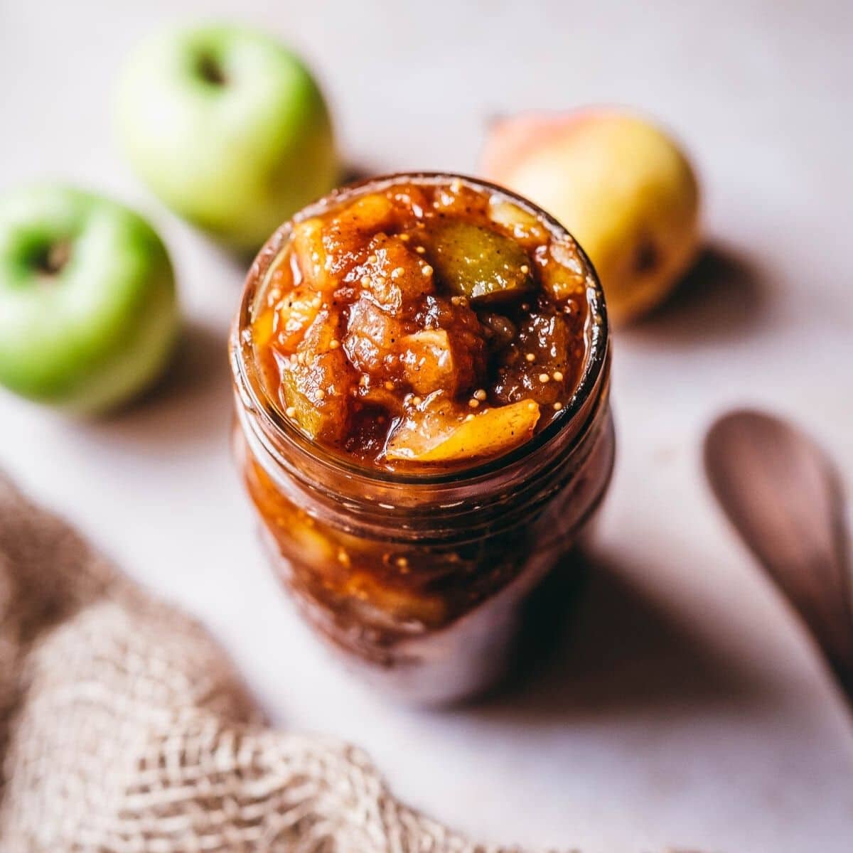 A glass jar filled with chunky apple chutney sits on a light surface, with two green apples and a yellow pear in the background—perfect for easy Christmas recipes and adding flavor to your stress-free Christmas table.