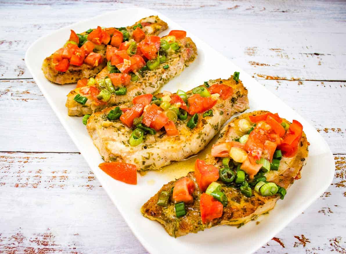 Three seasoned chicken breasts on a white rectangular plate, topped with chopped tomatoes and green onions&mdash;a perfect choice for quick meals on a rustic white wooden surface.
