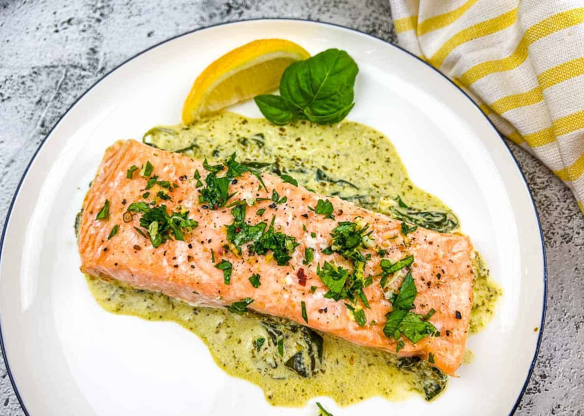 A piece of cooked salmon topped with chopped herbs sits on a creamy spinach sauce, garnished with a lemon wedge and basil, on a white plate&mdash;a perfect choice for quick dinner recipes.