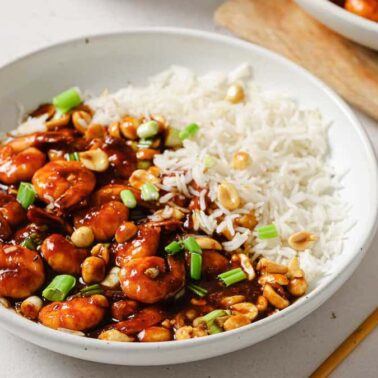 A bowl of white rice served with saucy shrimp, chopped green onions, and peanuts, placed on a white surface next to chopsticks—capturing true flavor harmony often found in Chinese recipes.