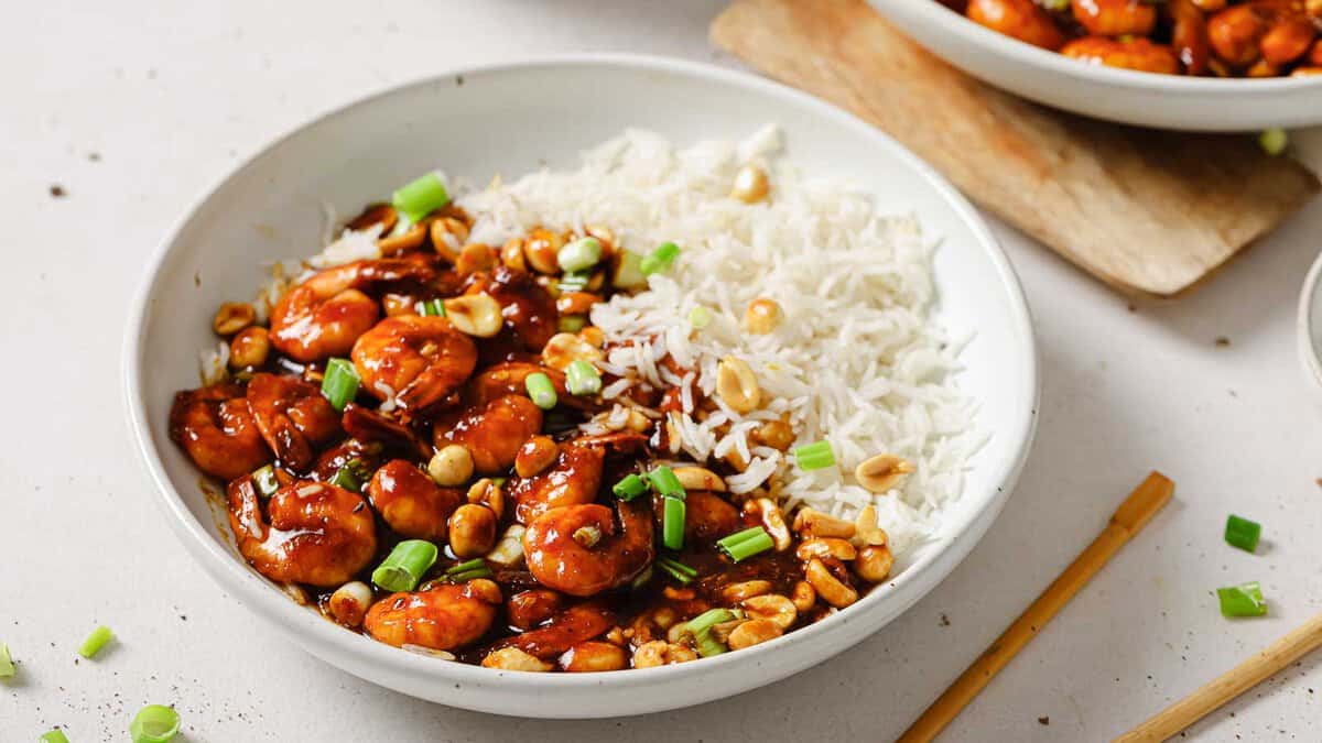 A bowl of white rice served with saucy shrimp, chopped green onions, and peanuts, placed on a white surface next to chopsticks—capturing true flavor harmony often found in Chinese recipes.