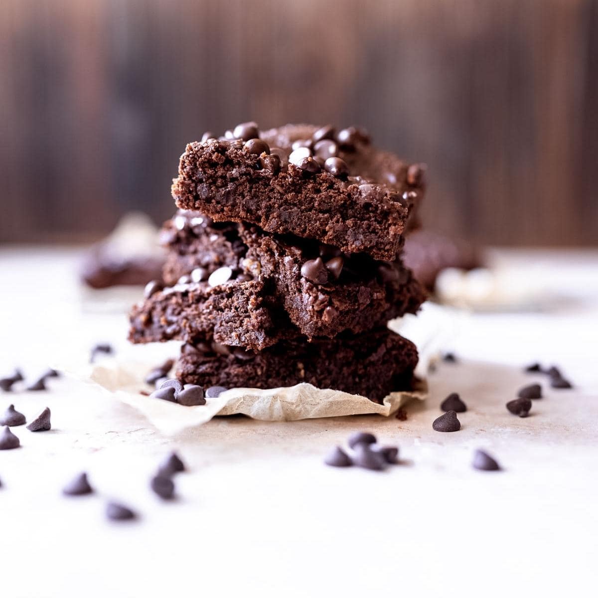 A stack of chocolate brownies with chocolate chips on top, placed on parchment paper, with chocolate chips scattered around&mdash;a decadent treat perfect for Gluten-Free Christmas Recipes.