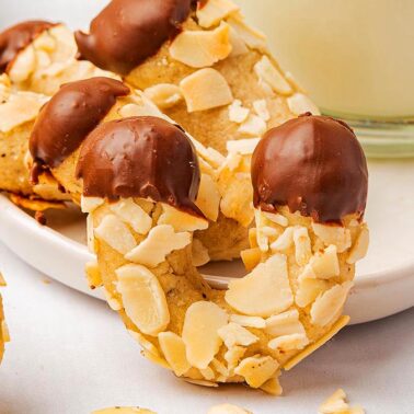 Curved cookies coated with almond slices and dipped in chocolate at both ends, placed on a white platter next to a glass of milk—perfect for your Gluten-Free Christmas Recipes collection.