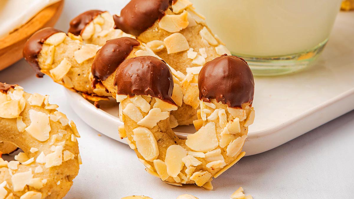 Curved cookies coated with almond slices and dipped in chocolate at both ends, placed on a white platter next to a glass of milk&mdash;perfect for your Gluten-Free Christmas Recipes collection.