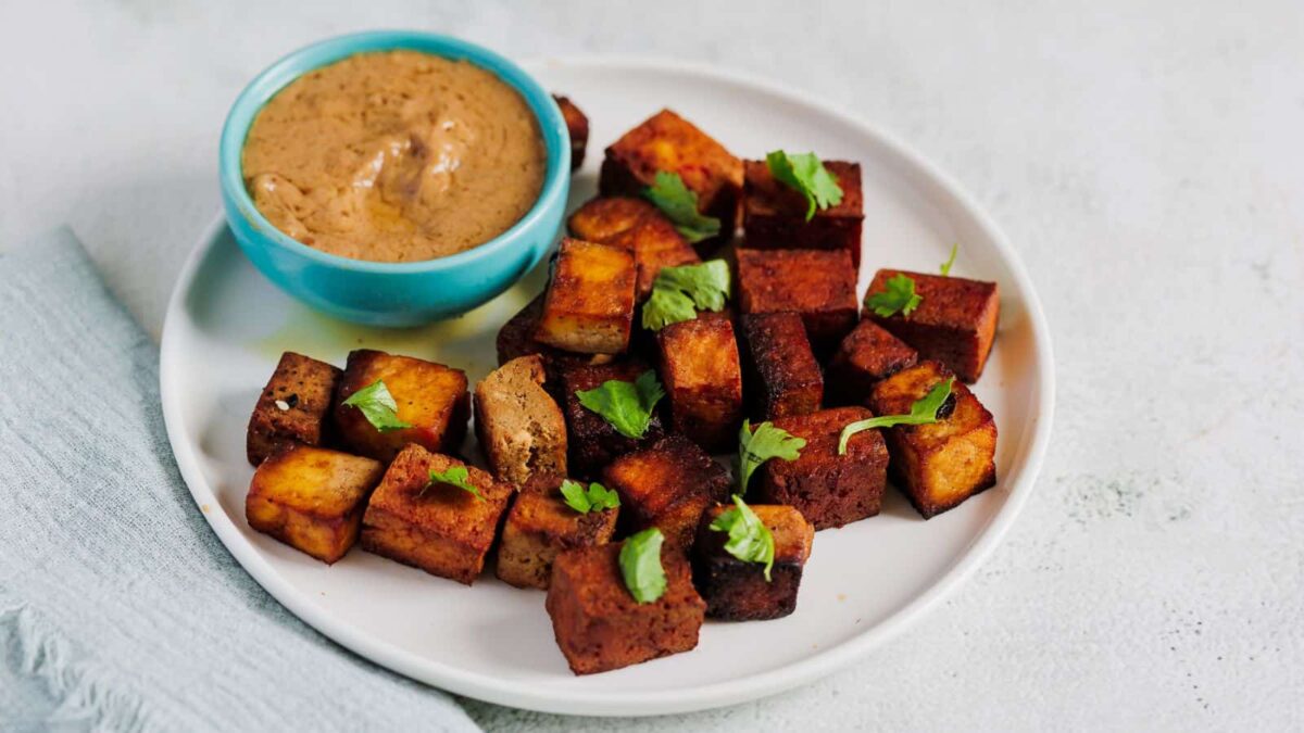 A white plate with crispy tofu cubes—an excellent source of plant protein—garnished with cilantro and served with a small bowl of dipping sauce.