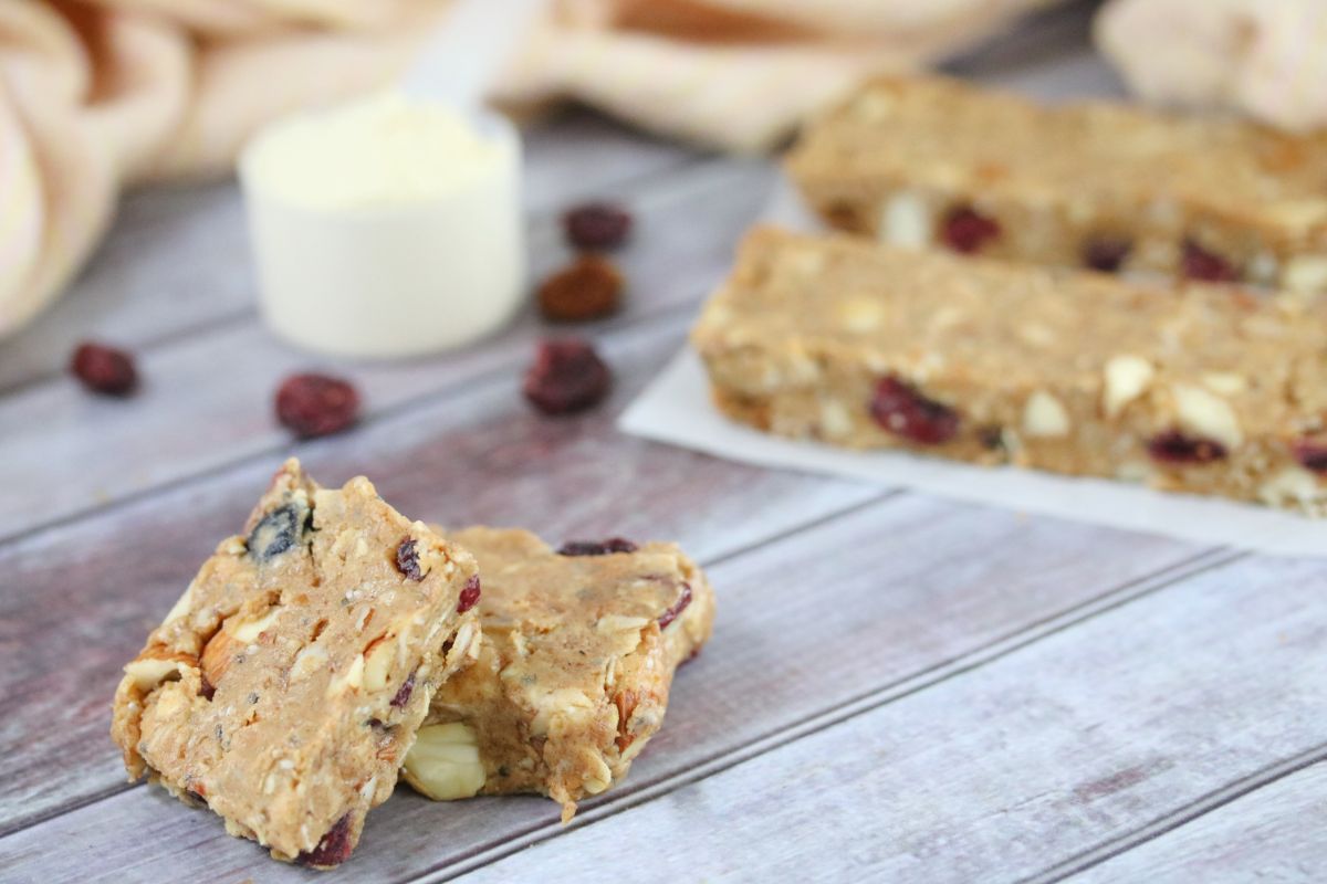 Two homemade protein bars with nuts and dried fruit are displayed on a wooden surface, perfect for healthy snacks that energize your day. A scoop of protein powder and more bars in the background hint at delicious protein snack recipes.