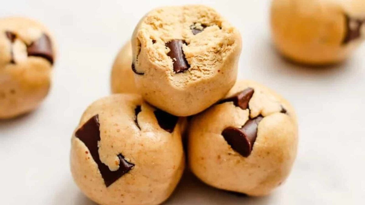 Four balls of raw cookie dough with visible chunks of chocolate are stacked on a light surface, perfect for healthy snacks that energize your day.