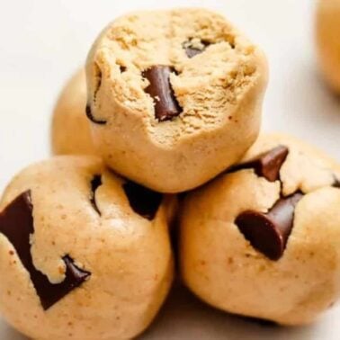 Four balls of raw cookie dough with visible chunks of chocolate are stacked on a light surface, perfect for healthy snacks that energize your day.
