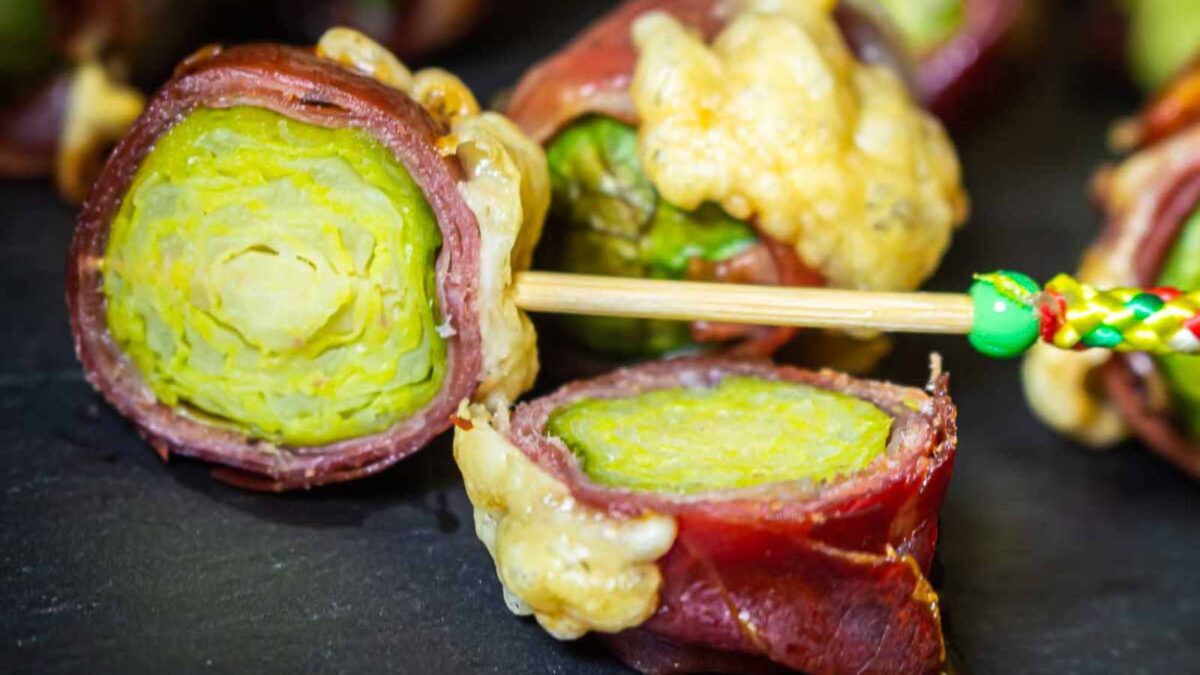Close-up of brussels sprouts wrapped in prosciutto with melted cheese, one piece held by a decorative toothpick—perfect for fans of bacon wrapped recipes and smoky-salty sweet recipes.