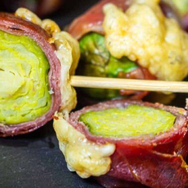 Close-up of brussels sprouts wrapped in prosciutto with melted cheese, one piece held by a decorative toothpick—perfect for fans of bacon wrapped recipes and smoky-salty sweet recipes.