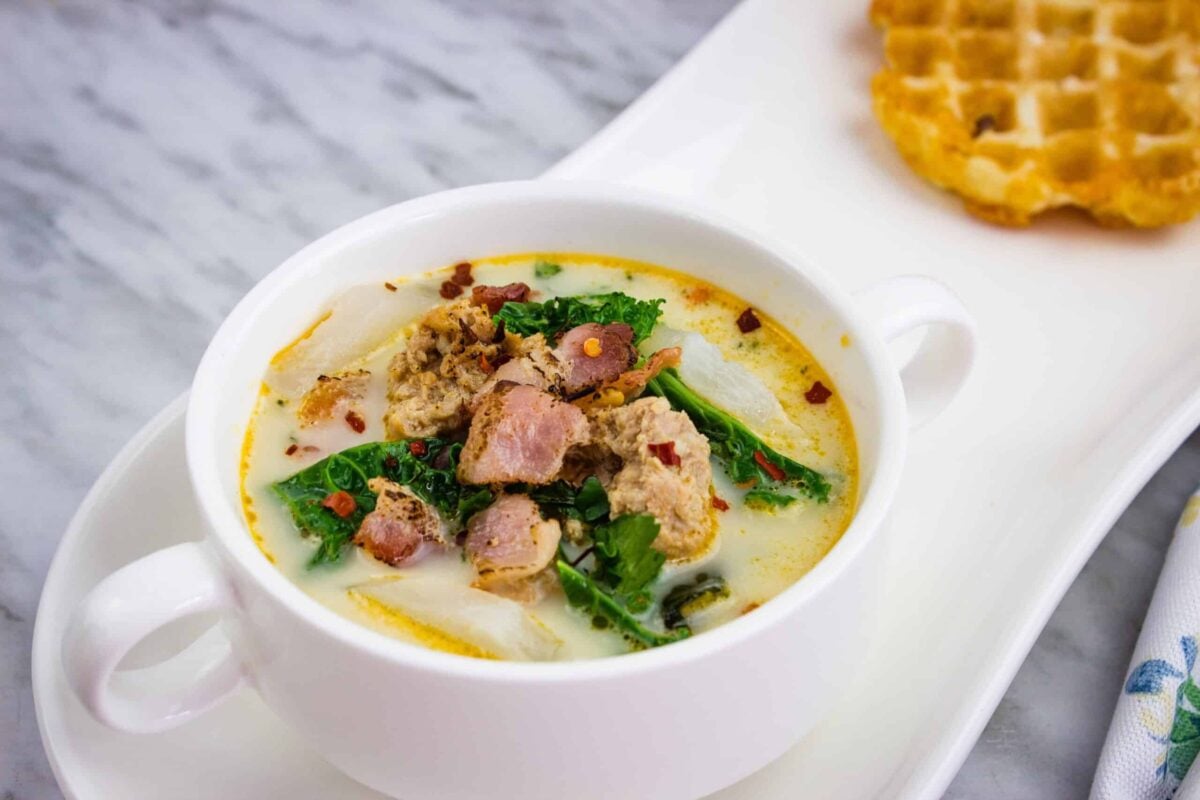 A white bowl of creamy, low-carb soup with sausage, kale, and bacon bits, served on a white plate with a waffle on the side.