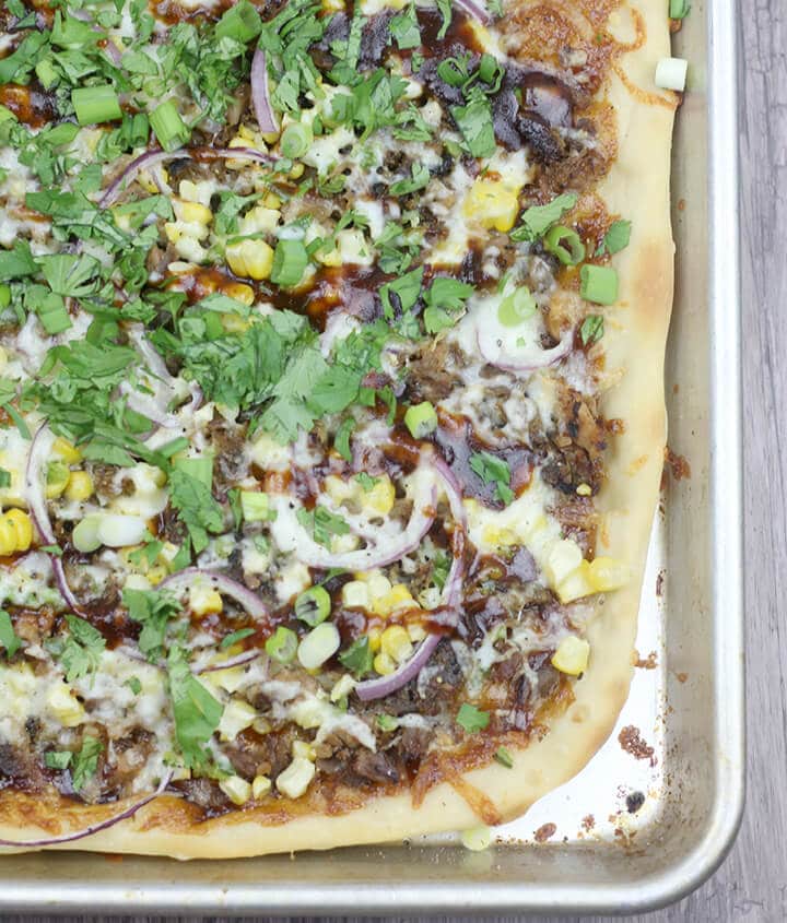 Rectangular homemade pizza with melted cheese, red onion, corn, green onions, cilantro, and sauce on a baking sheet—a delicious twist on smoked recipes with a hint of smoked flavor in every bite.