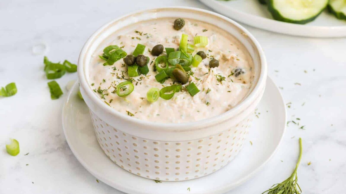 A ceramic bowl filled with a creamy dip boasting smoked flavor, garnished with chopped green onions, capers, and herbs, placed on a small white plate.