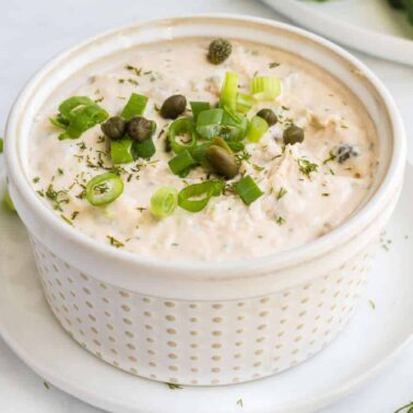 A ceramic bowl filled with a creamy dip boasting smoked flavor, garnished with chopped green onions, capers, and herbs, placed on a small white plate.