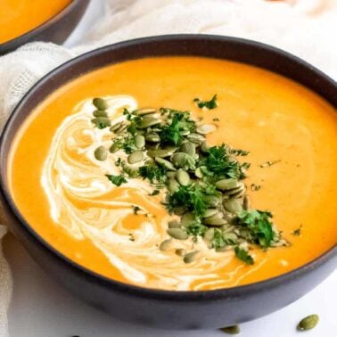 A bowl of creamy orange soup with a hint of curry flavor, topped with a swirl of cream, chopped parsley, and pumpkin seeds, sits on a white surface next to a light-colored cloth.