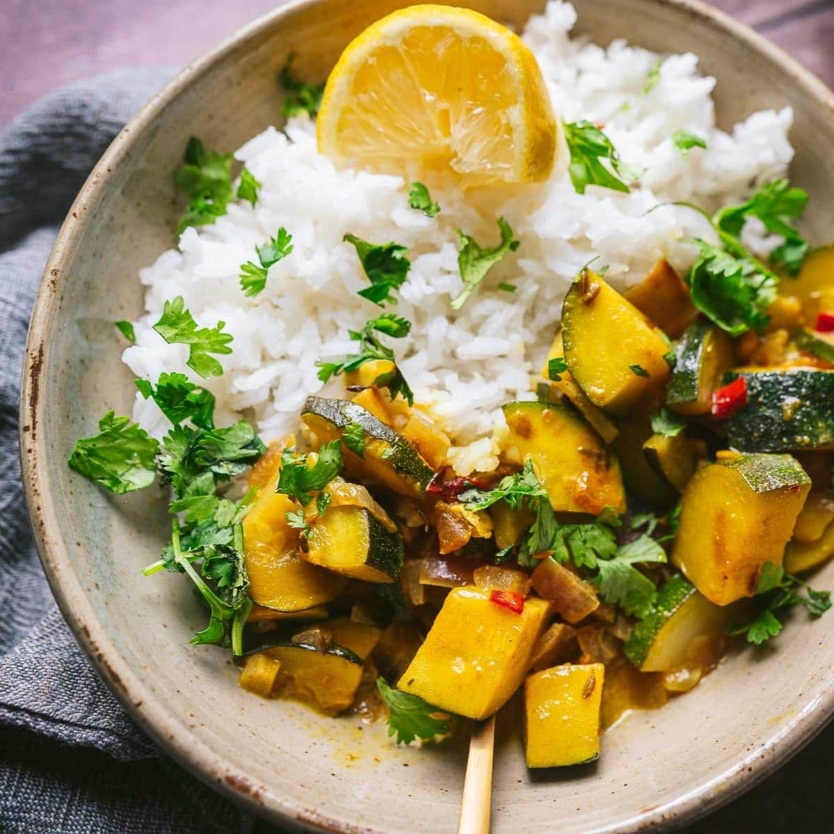 A bowl of white rice topped with zucchini curry, garnished with fresh cilantro and a lemon wedge—perfect for those who love trying new curry recipes.