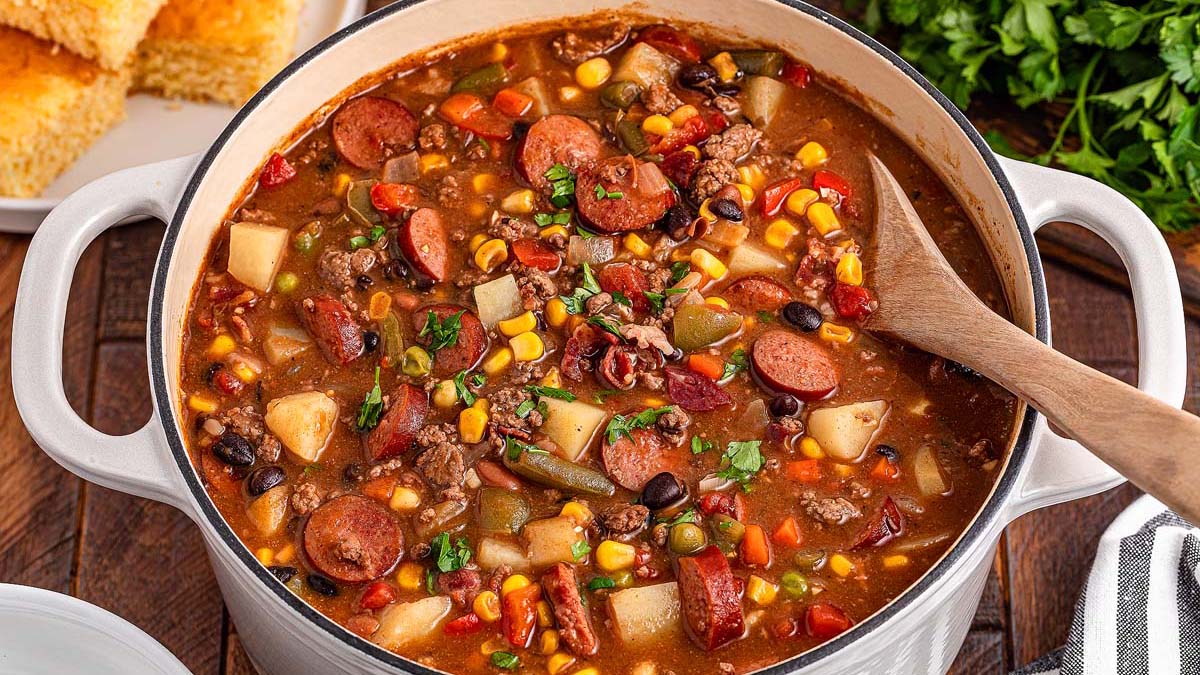 A pot of hearty stew with soulful flavor, packed with sausage, ground beef, potatoes, corn, beans, tomatoes, and mixed vegetables, served with a wooden spoon and cornbread on the side.