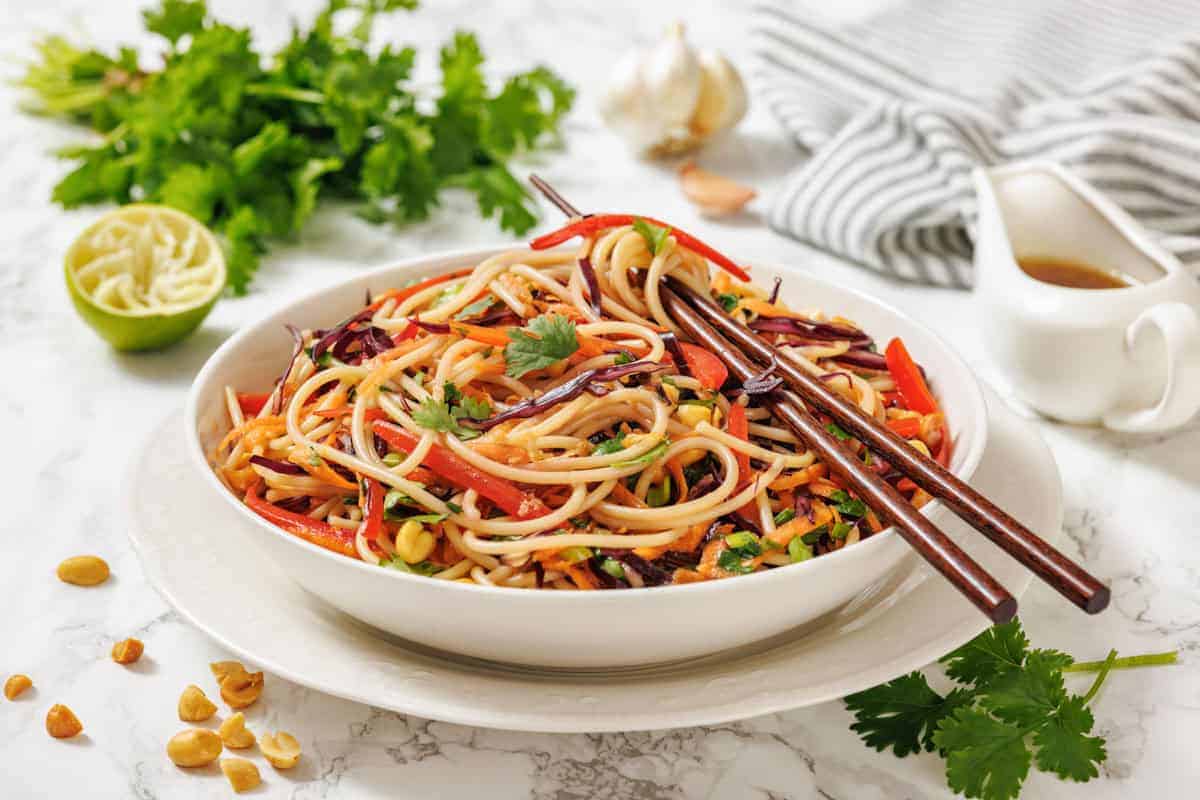 A bowl of Asian-inspired noodle salad with vegetables and chopsticks, garnished with cilantro, on a white plate—with sauce, lime, peanuts, and herbs nearby. Perfect for those seeking fresh dressing ideas to brighten salads and elevate their salad bowls.